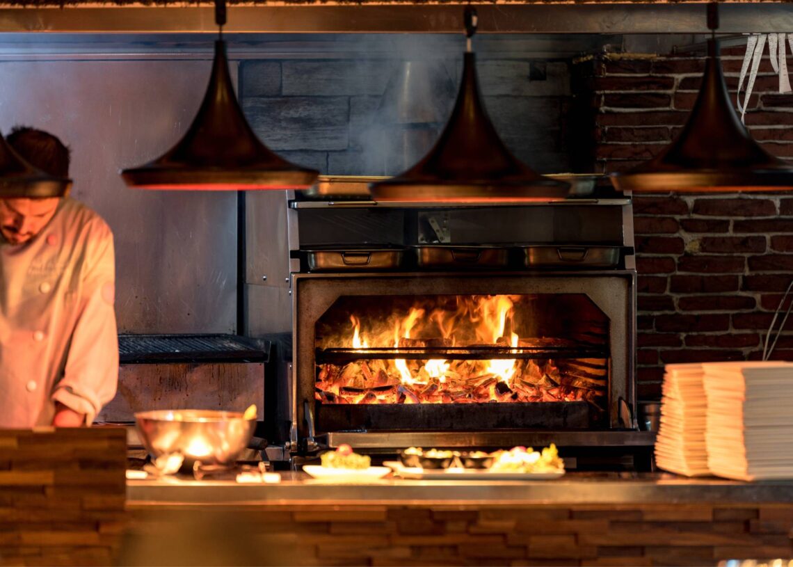 man in a chef outfit working at a pizzeria as a wood-fired oven burns in the background with ingredients in the foreground