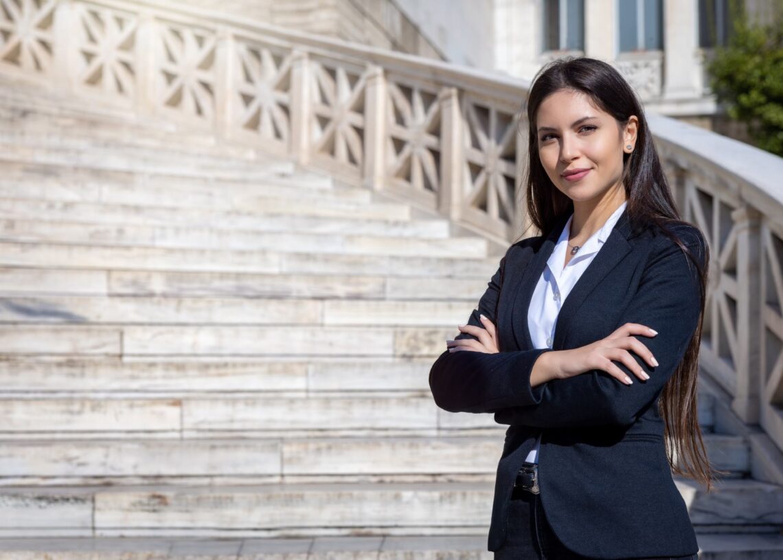 A professional young woman with a confident demeanor standing with her arms crossed in front of a white courthouse staircase.