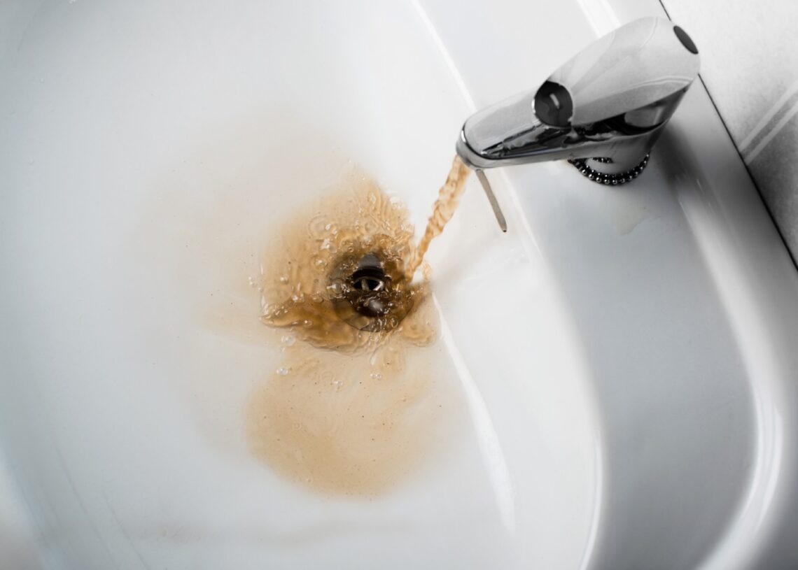A bathroom porcelain sink with a silver faucet running a stream of brown stained water down the drain.