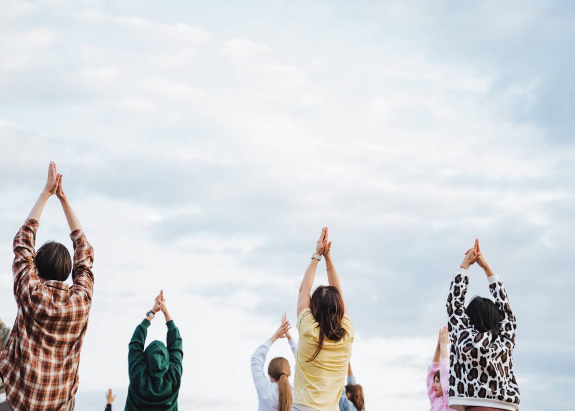 A group of people stands outside with their arms in the air. They hold their hands together, making a pyramid shape.