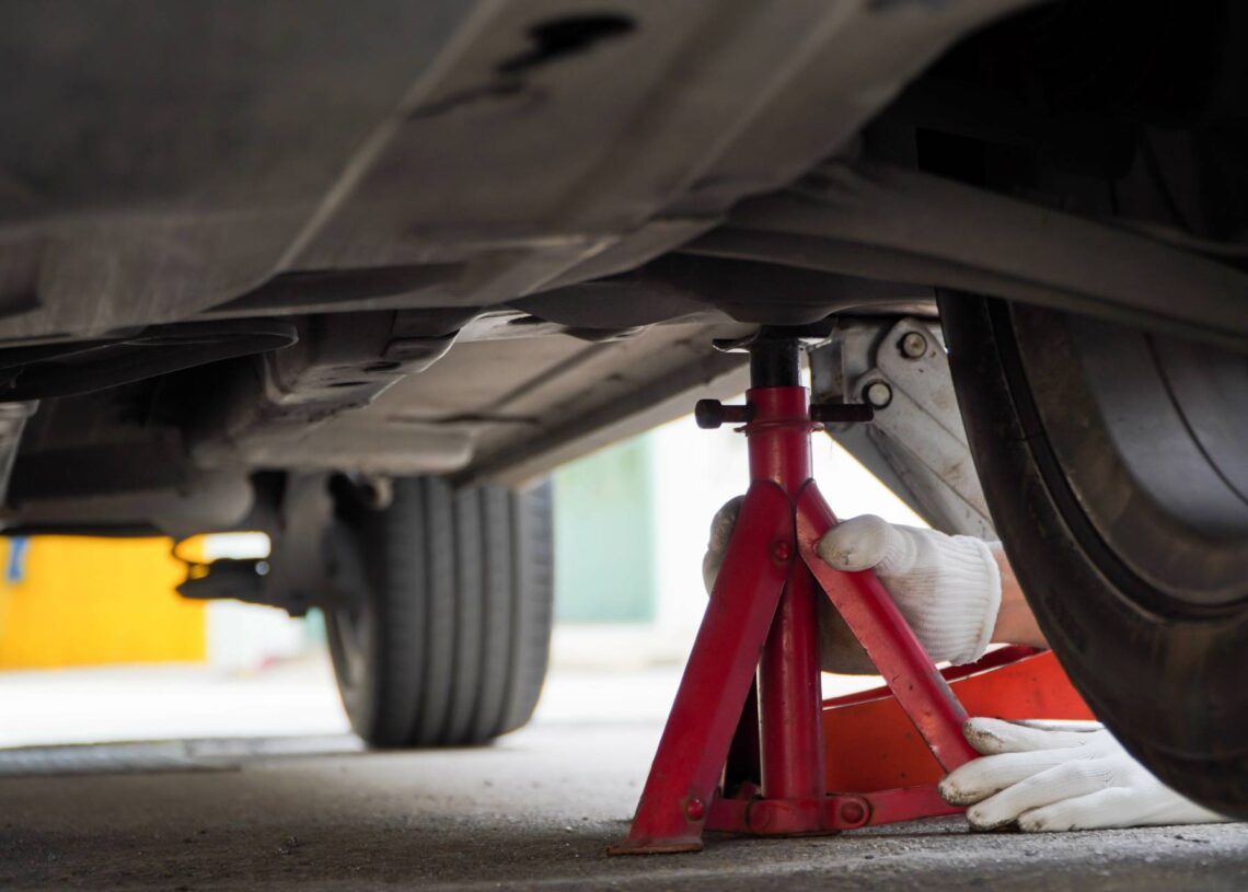 A person's hands place a red jack stand underneath a vehicle, near the rear tire. The person is wearing white gloves.