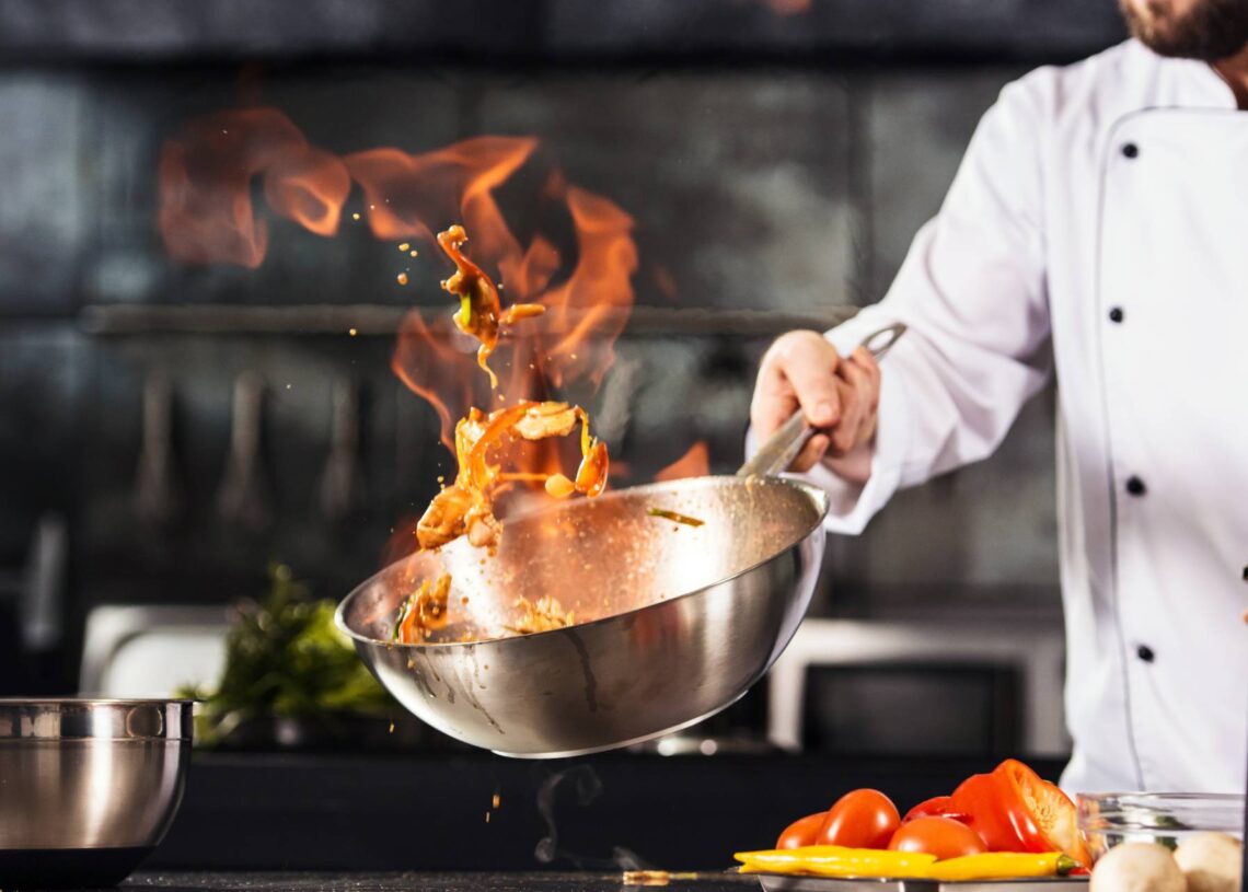 A chef moves a wok around to cook food, the flames rising from the wok as food is caught midair. Cut ingredients are on the counter beside the chef.