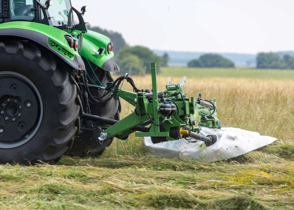 A mower attachment on a large green tractor cuts tall grass in a rural agricultural field, bordered by trees.
