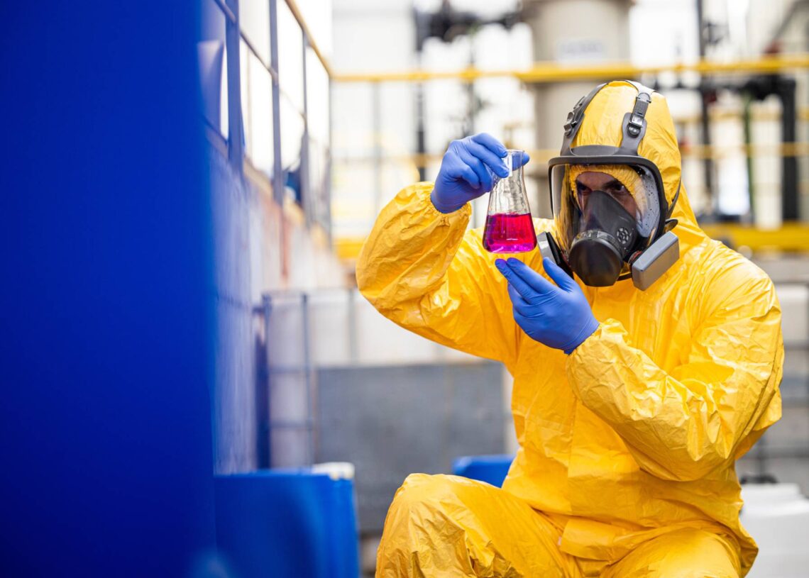 A male employee wearing a yellow hazmat suit and a gas mask is looking at a beaker filled with a red liquid.