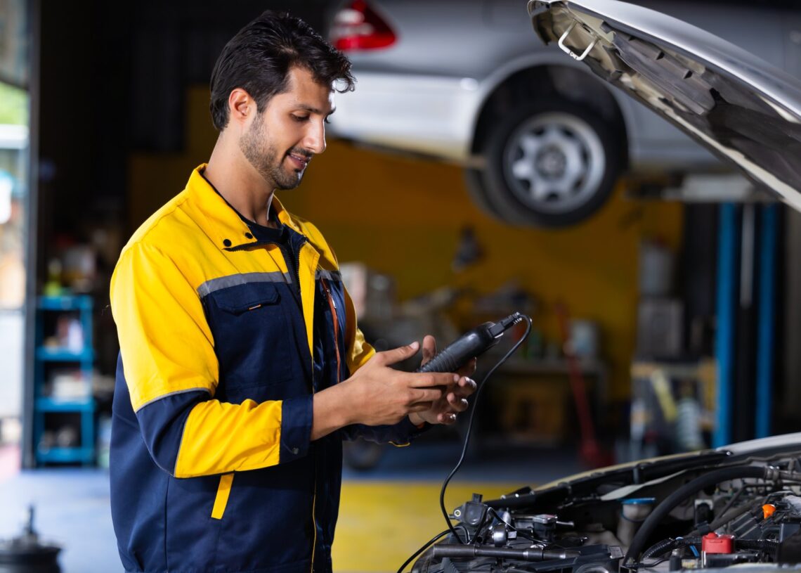 A man wearing a blue and yellow jacket standing in front of an open car hood. He is checking the battery with a handheld device.