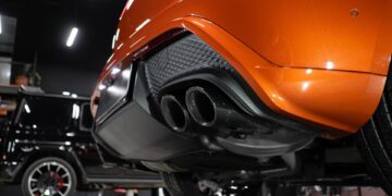 A close-up of a modern and luxury sports car exhaust pipe. The vehicle is an orange color and is parked in a garage.