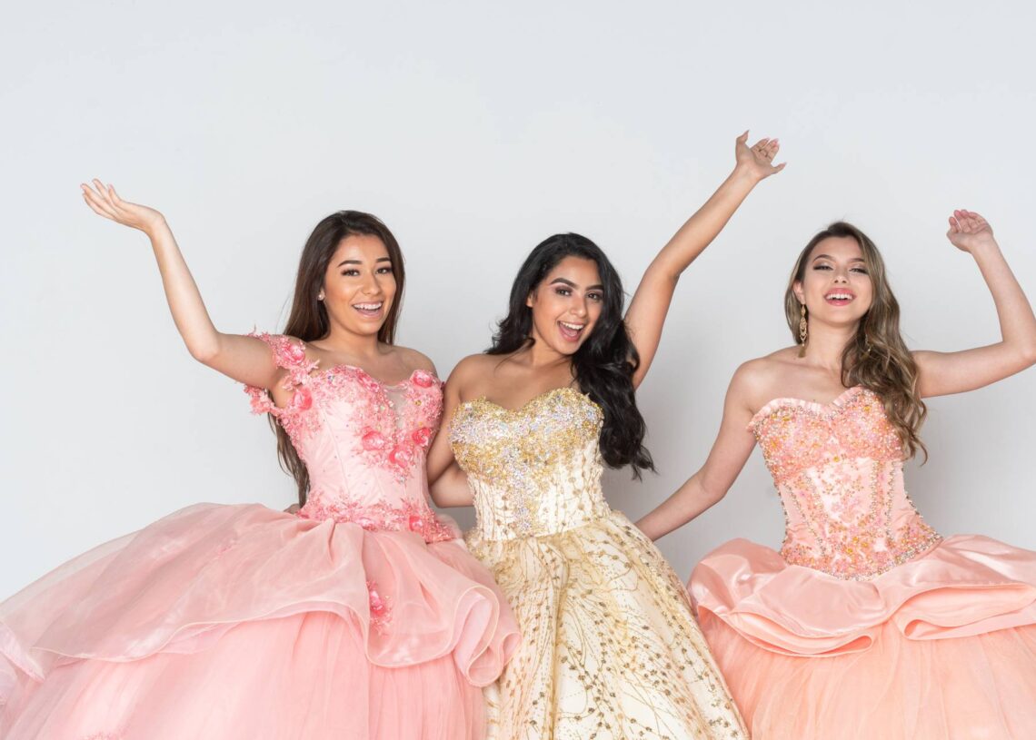 Three girls in quinceañera dresses stand beside one another. The one in middle has a gold dress while the other two wear pink.