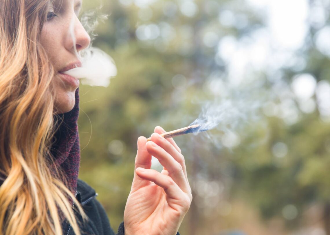 A close-up side view of a woman smoking a cannabis joint. She has brown and blonde hair and wears a jacket.