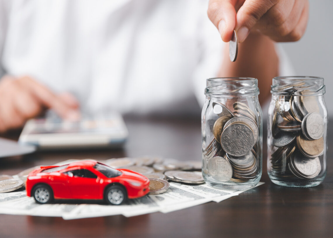 Someone dropping coins into small glass jars next to a small red toy car that rests on a brown table.