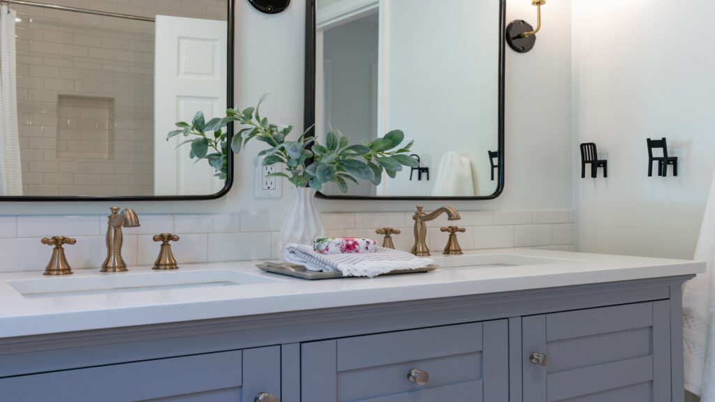 A gray bathroom vanity with two sinks, brass hardware, white granite countertops, and two mirrors above each sink.