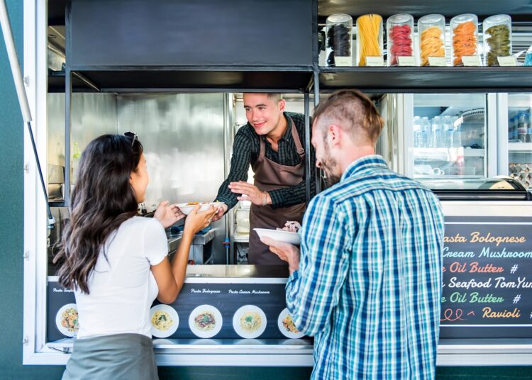 A woman is accepting food from a man working on a food truck. A different man stands beside the woman holding food.