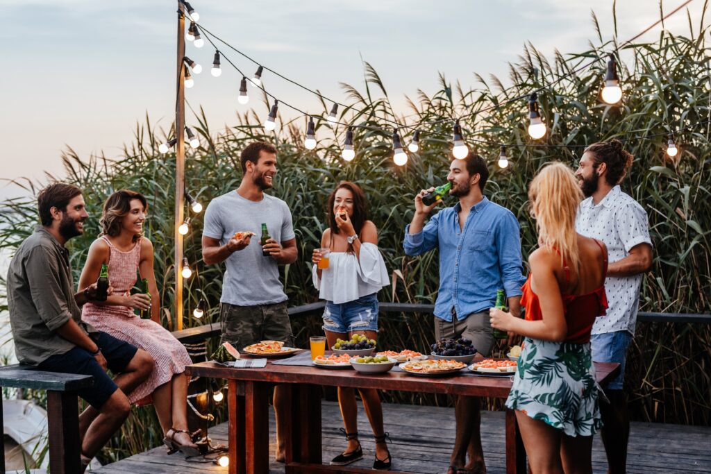 A group of friends stand around a table, smiling, laughing, and drinking. There are plates of food on the table.