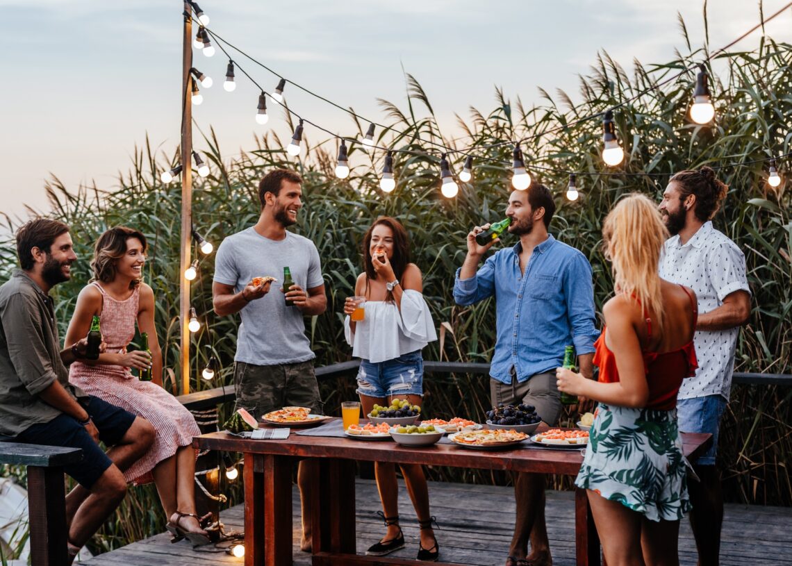 A group of friends stand around a table, smiling, laughing, and drinking. There are plates of food on the table.
