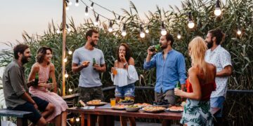 A group of friends stand around a table, smiling, laughing, and drinking. There are plates of food on the table.