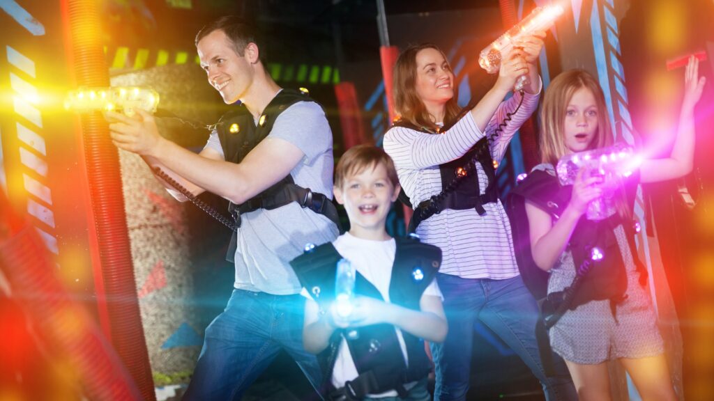 A family of two parents and a young boy and girl are playing laser tag with vests and laser blasters in an arena.