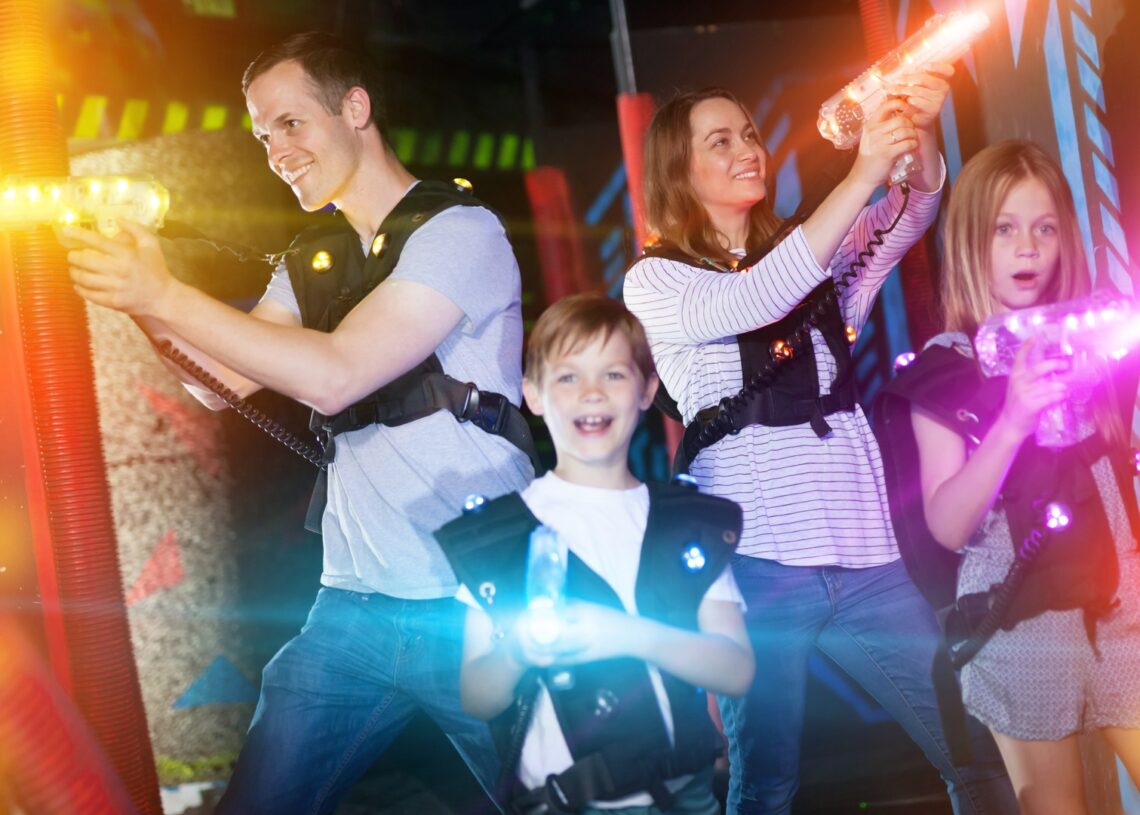 A family of two parents and a young boy and girl are playing laser tag with vests and laser blasters in an arena.