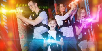 A family of two parents and a young boy and girl are playing laser tag with vests and laser blasters in an arena.