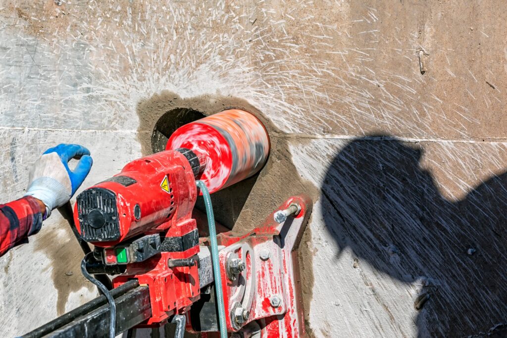 A worker using a red drilling machine to core into a large concrete wall with another worker's shadow nearby.