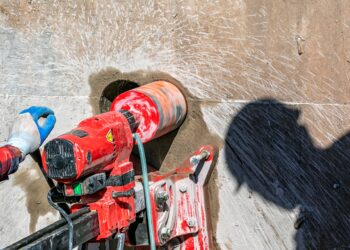 A worker using a red drilling machine to core into a large concrete wall with another worker's shadow nearby.