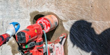 A worker using a red drilling machine to core into a large concrete wall with another worker's shadow nearby.