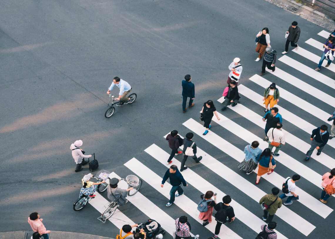 An aerial view shows several people walking across a crosswalk on a road. Some people are riding bikes.