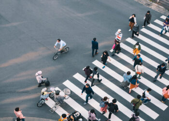 An aerial view shows several people walking across a crosswalk on a road. Some people are riding bikes.