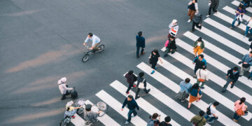 An aerial view shows several people walking across a crosswalk on a road. Some people are riding bikes.