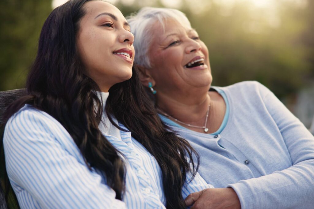 A happy older mother and woman sitting on a bench, enjoying nature and sharing a laugh with each other.