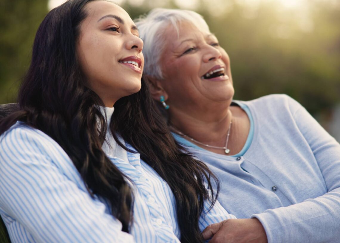 A happy older mother and woman sitting on a bench, enjoying nature and sharing a laugh with each other.