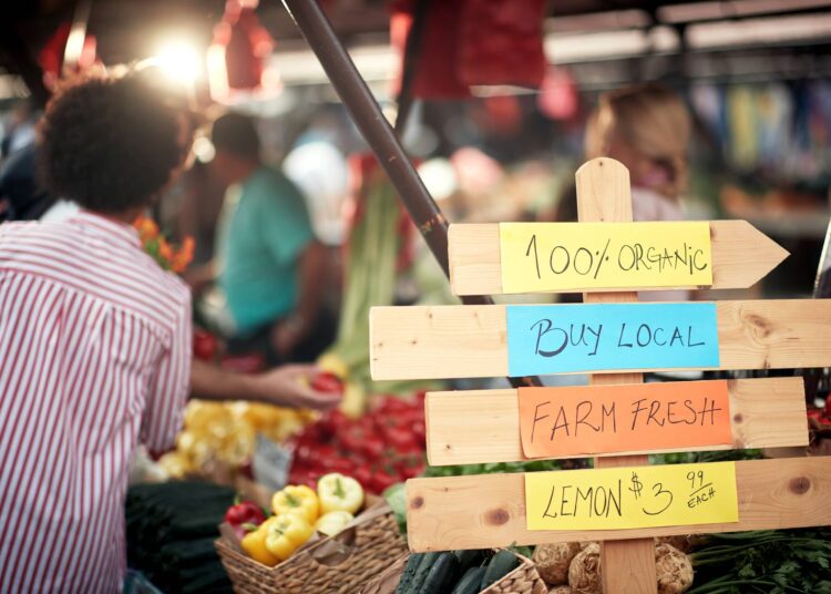 A wooden sign contains handwritten notes that read “100% organic, buy local, farm fresh, lemons $3.99.”