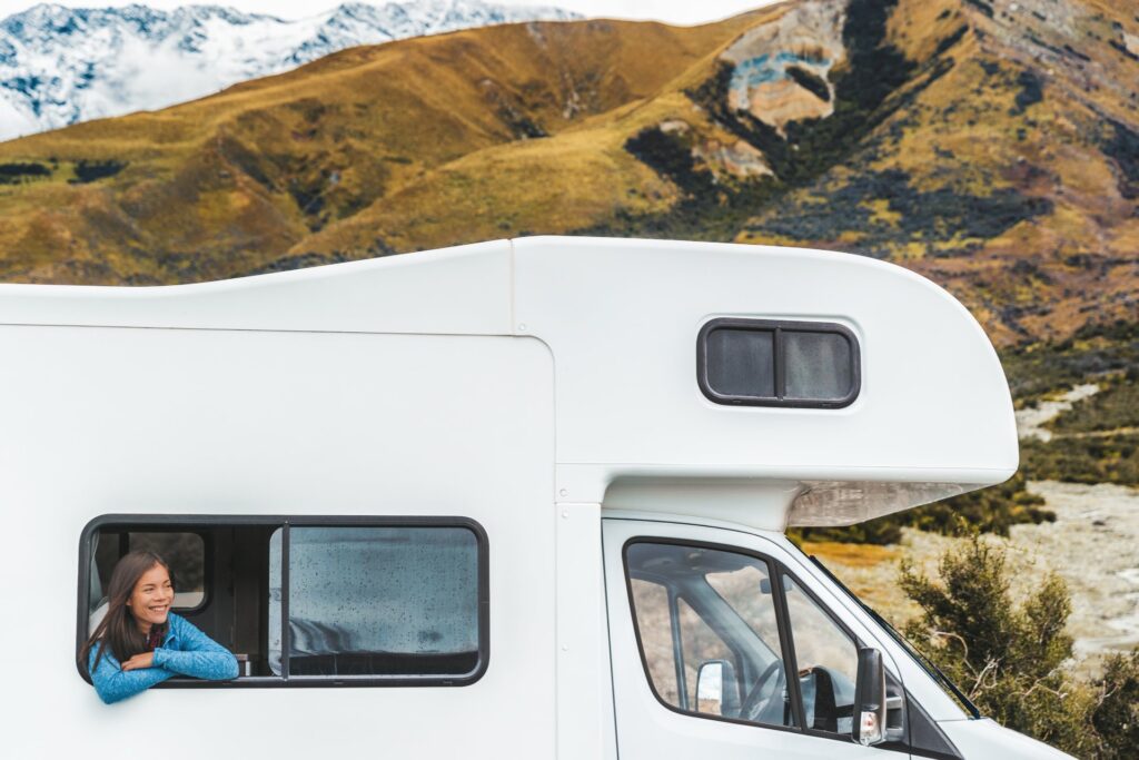 A woman smiles, folds her arms, and leans out of her RV's window. Large mountains are in the background.