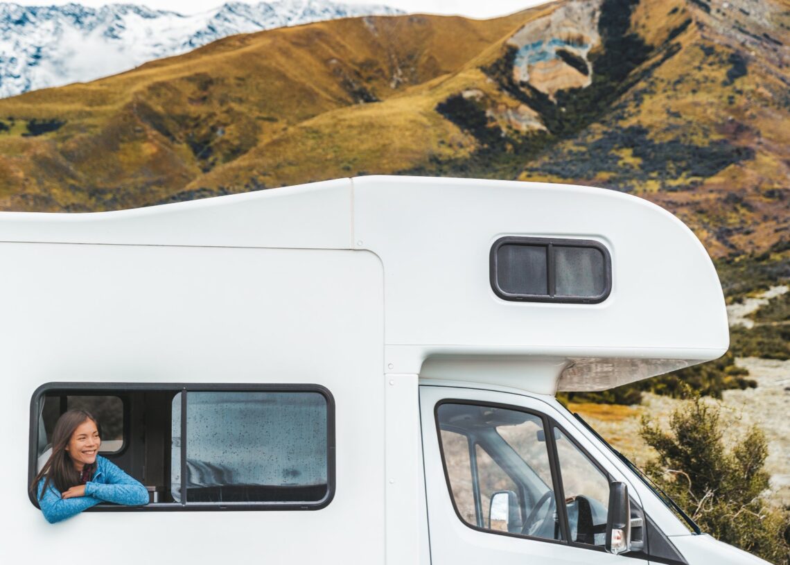 A woman smiles, folds her arms, and leans out of her RV's window. Large mountains are in the background.