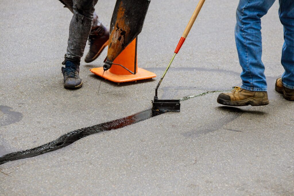 Two individuals in jeans using tools to apply black sealant to repair a long crack in an asphalt surface.