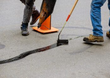 Two individuals in jeans using tools to apply black sealant to repair a long crack in an asphalt surface.