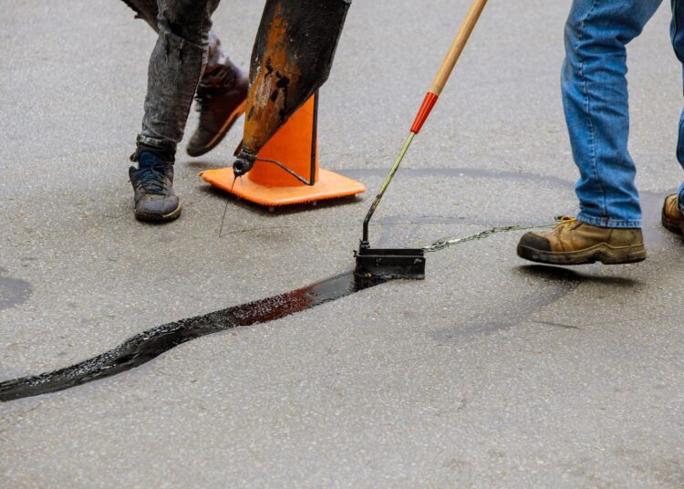 Two individuals in jeans using tools to apply black sealant to repair a long crack in an asphalt surface.