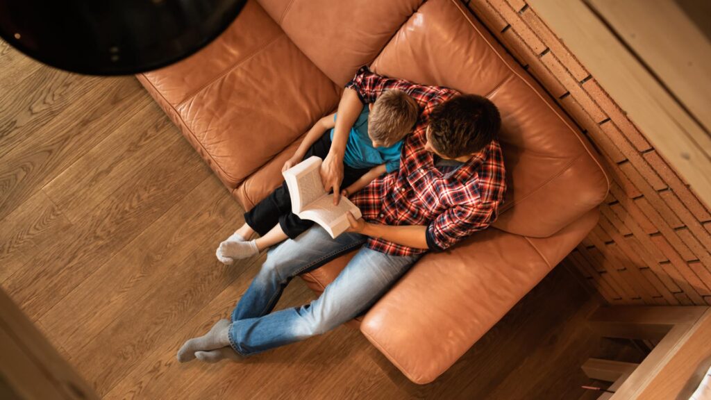 An aerial view shows a father and son sitting on a brown leather couch, reading the Bible. The father has an arm around his son.