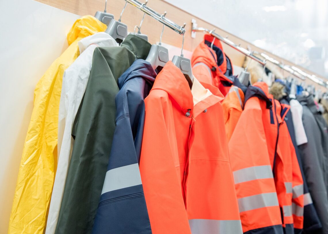 Different blue-collar workplace safety clothing tops hang on a metal rack inside a brightly-lit store.