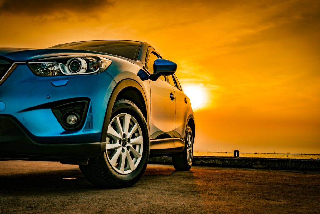 A sleek blue sedan parked on concrete. Behind the vehicle is a view of the sky with a setting sun over a body of water.
