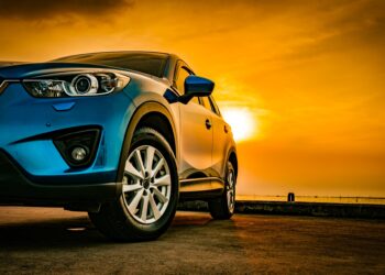 A sleek blue sedan parked on concrete. Behind the vehicle is a view of the sky with a setting sun over a body of water.
