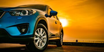 A sleek blue sedan parked on concrete. Behind the vehicle is a view of the sky with a setting sun over a body of water.