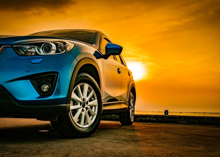 A sleek blue sedan parked on concrete. Behind the vehicle is a view of the sky with a setting sun over a body of water.