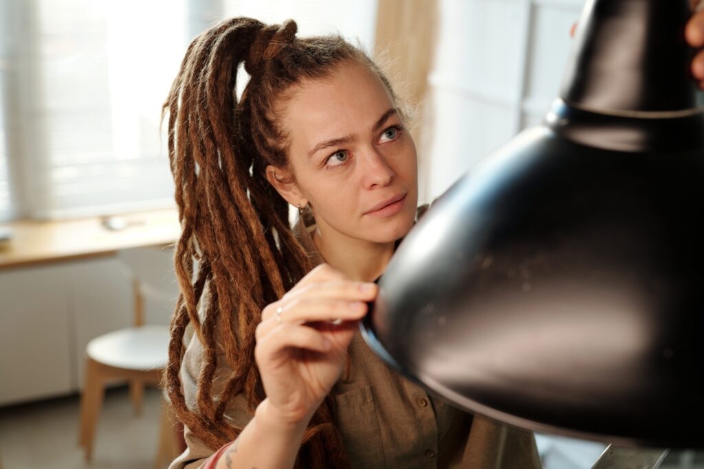 A woman with long dreadlocks pulled back in a ponytail examining a black overhead lamp inside a well-lit room.