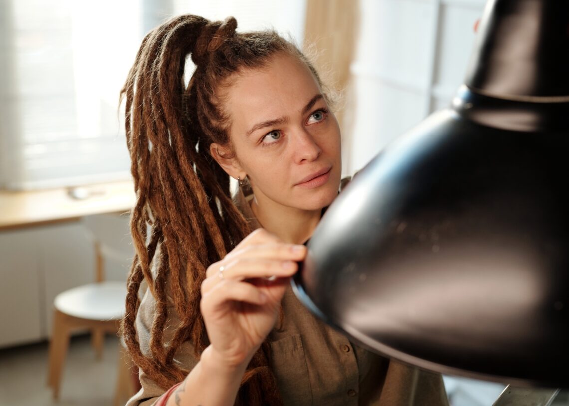 A woman with long dreadlocks pulled back in a ponytail examining a black overhead lamp inside a well-lit room.