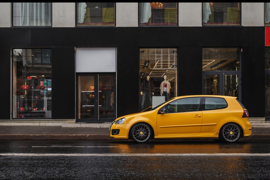 A small yellow car is parked in front of a retail store in a city. The road around them is clear of other vehicles.