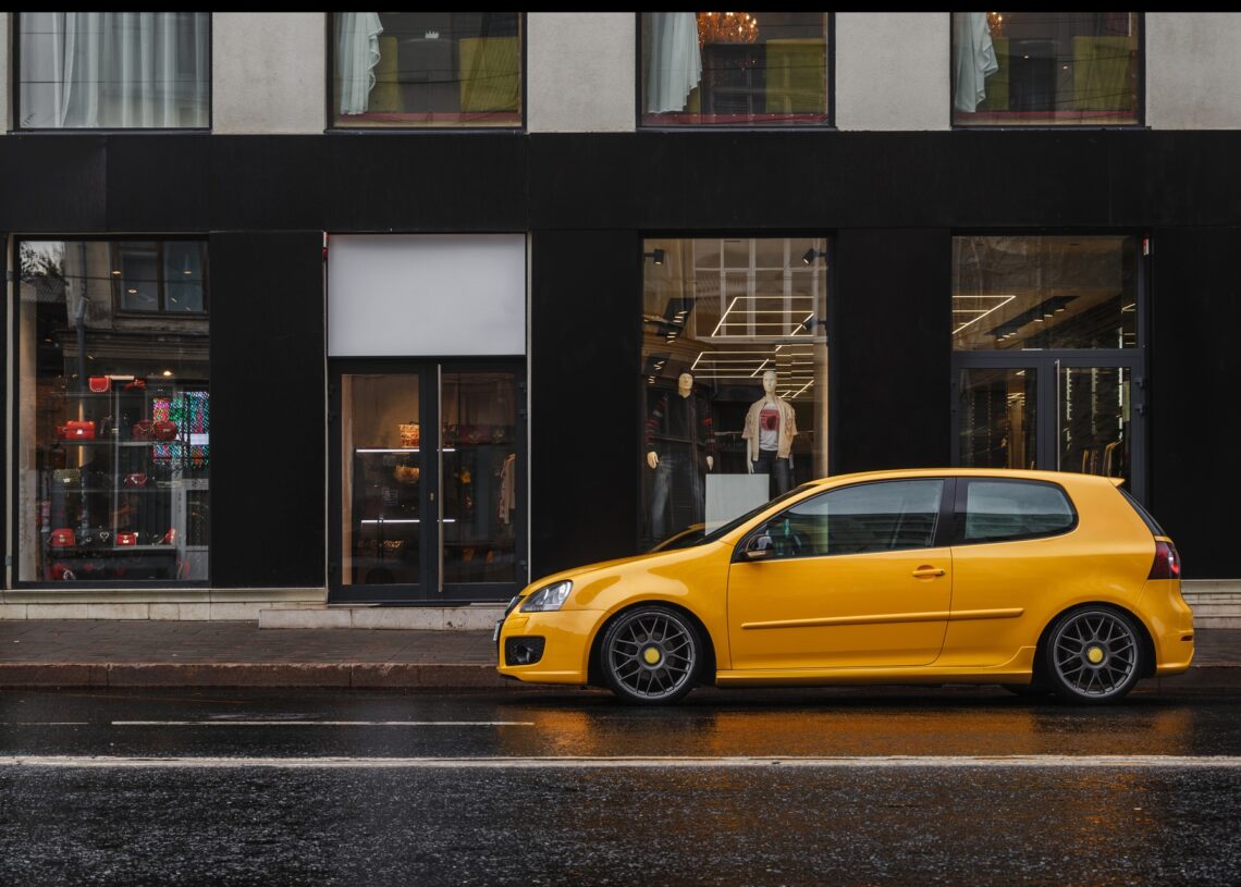 A small yellow car is parked in front of a retail store in a city. The road around them is clear of other vehicles.