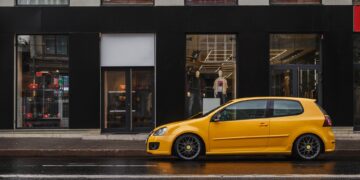 A small yellow car is parked in front of a retail store in a city. The road around them is clear of other vehicles.
