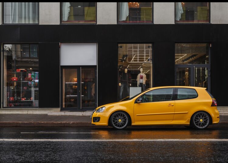 A small yellow car is parked in front of a retail store in a city. The road around them is clear of other vehicles.