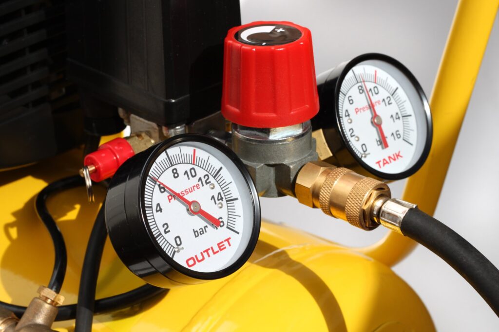 A yellow air compressor up-close with two gauges and a red turn dial. The air hoses are black around the gauges.