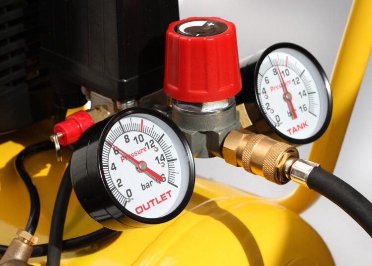 A yellow air compressor up-close with two gauges and a red turn dial. The air hoses are black around the gauges.