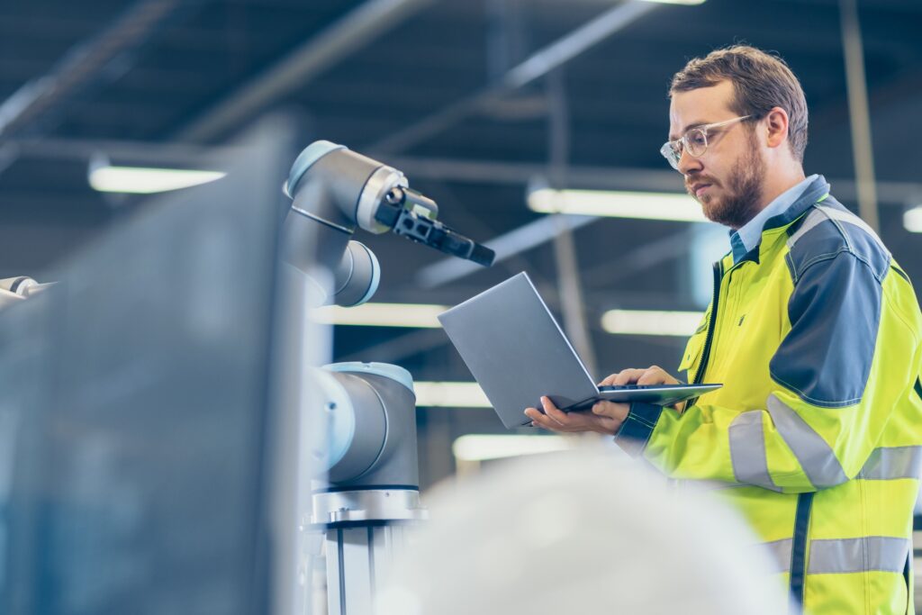 A factory employee wears a yellow safety vest as he works on a laptop in front of a robotic machine.